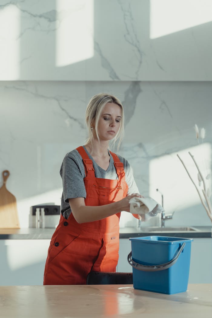 about-01 A woman wearing orange overalls cleans a kitchen counter with a rag and pail in a modern kitchen.